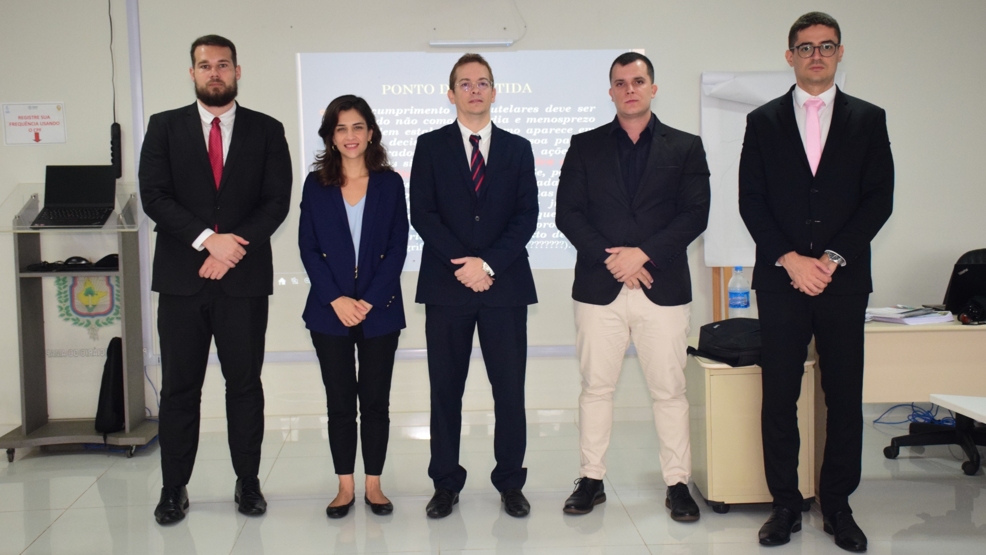  Juiz Diogo Moura de Araújo e mais quatro novos Juízes em pé posando para a foto em uma sala de aula e ao fundo um Datashow apresentado.